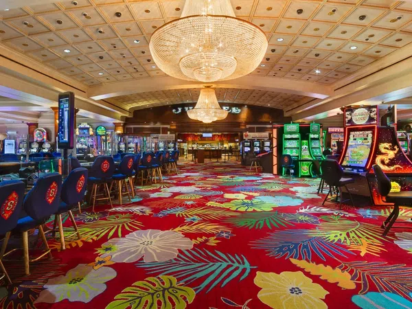 Colorful casino interior with floral carpet, gaming machines, blue chairs, and large chandeliers overhead