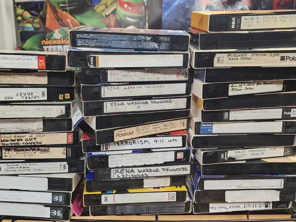 Stack of vintage VHS tapes with handwritten labels on a cluttered desk surrounded by posters and household items.