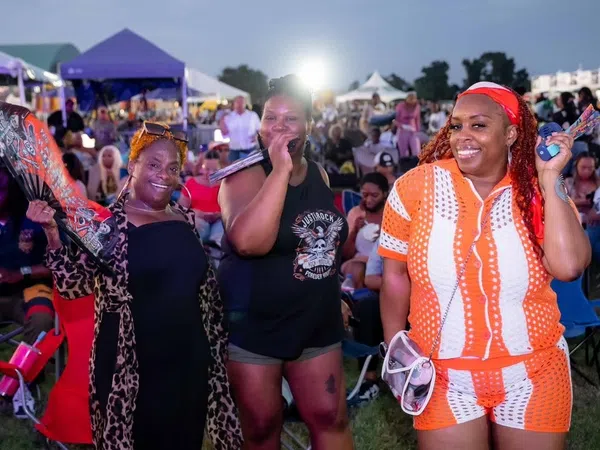 Three smiling women at an outdoor event with tents and crowd in the background during evening.