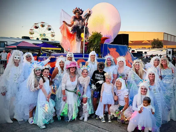 Group of people in white costumes and wings posing outdoors at a festival with a ferris wheel and colorful float.