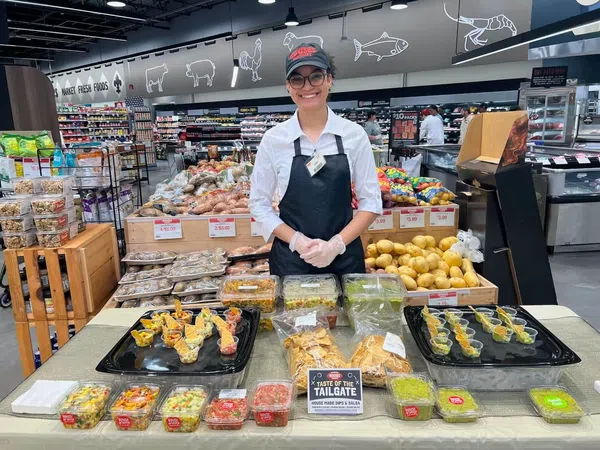 Smiling store employee presenting various homemade dips and salsas at a grocery store sampling table.