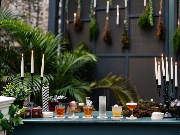 Outdoor bar setup with assorted cocktails, lit candles, greenery, and dried herbs hanging on a dark backdrop.