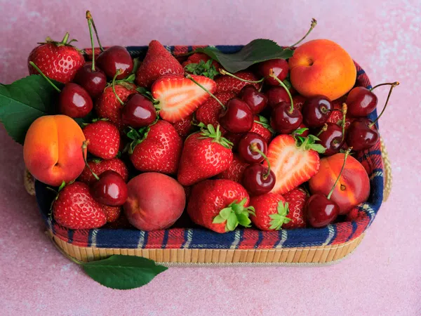 Basket filled with fresh strawberries, cherries, peaches, and apricots on a pink surface