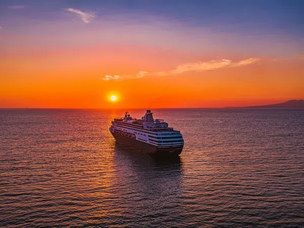 Large cruise ship sailing on calm ocean waters during vibrant orange sunset with clear sky.