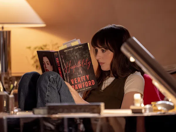 Woman reading the book Hospitality by Verity Crawford in a cozy, warmly lit room with a table lamp.