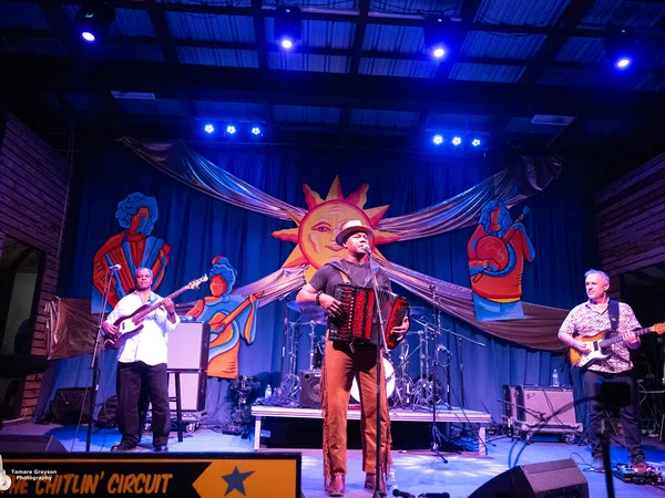 Live band performing on stage with accordion, guitar, and bass under bright lights and colorful sun backdrop