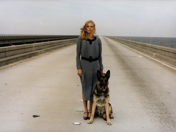 Woman in gray dress stands with German Shepherd on empty bridge over water under cloudy sky.