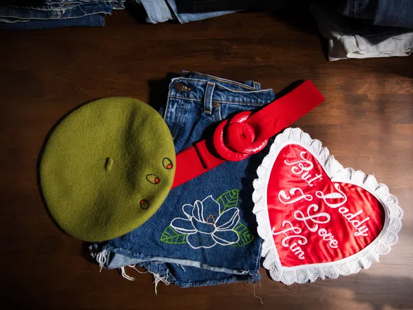 Green beret, denim shorts with floral embroidery, red belt, and heart-shaped pillow with love message on wooden surface.