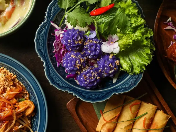 Colorful Thai dishes including purple flower-shaped dumplings, spring rolls, pad thai, and curry on ornate plates.