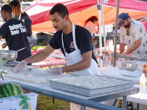 Man wearing apron preparing dough at outdoor food stall with trays of dough and tents in background