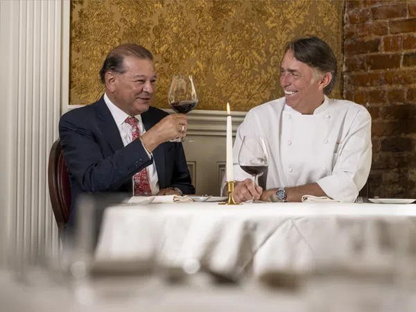 Two men smiling and toasting with red wine in an elegant dining room with gold wallpaper and exposed brick.