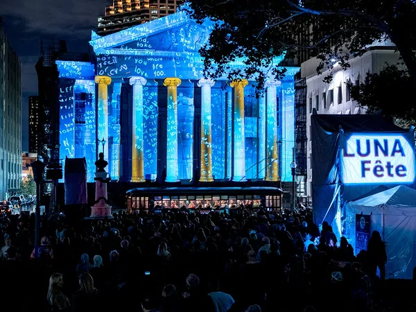 Crowd at night watching blue and gold light projections on a classical building during Luna Fête festival.