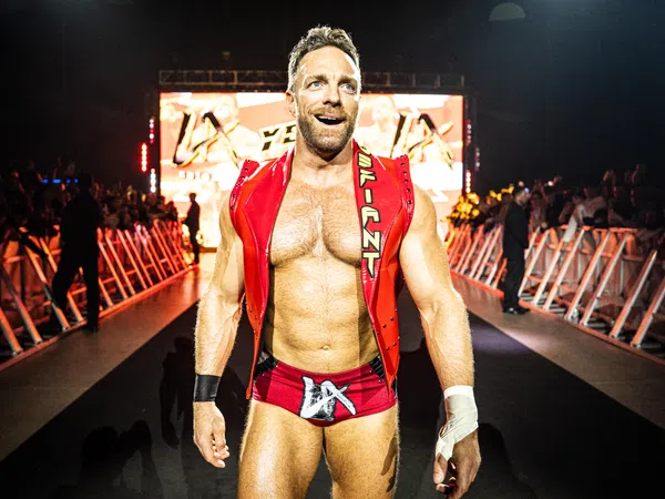 Muscular male wrestler in red trunks and vest walking confidently toward ring with audience and lights behind him