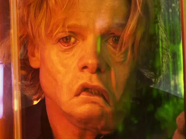 Close-up of a distressed man with blonde hair looking through a glass panel illuminated by orange and green light.