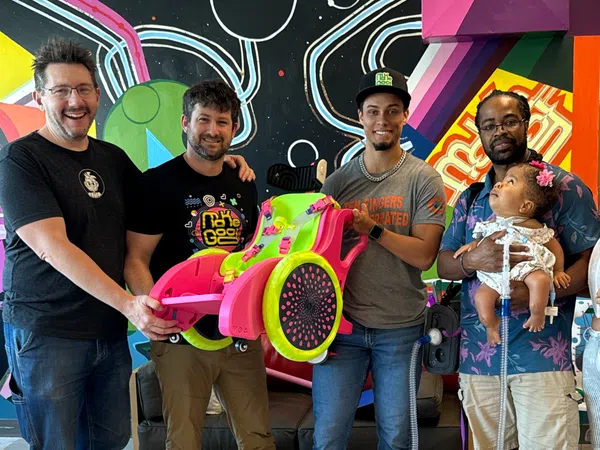 Group of five people smiling indoors with colorful wall art, holding a bright pink and green custom wheelchair.