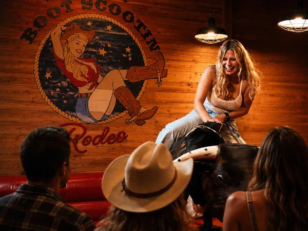 Young woman smiling while riding a mechanical bull in a wooden rodeo-themed bar with audience watching.