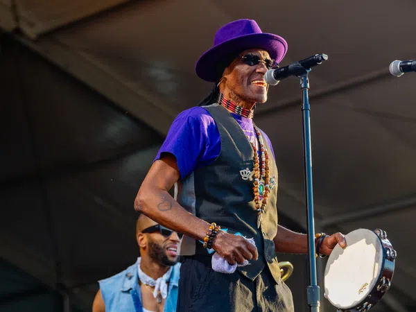 Musician in purple hat and vest singing into microphone while playing tambourine on stage at outdoor event.