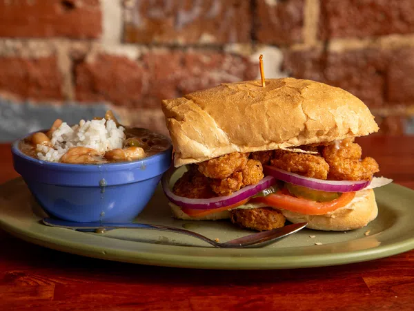 Crispy fried shrimp po' boy sandwich with lettuce, tomato, onion, served with a side of rice and beans in a blue bowl.