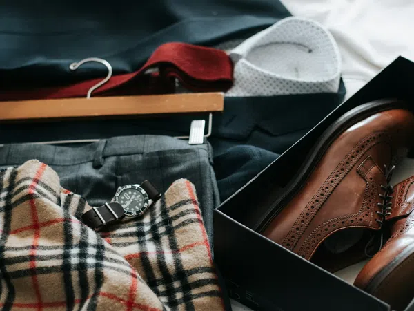 Flat lay of men's brown dress shoes in a box, plaid scarf, gray trousers, watch, and a white shirt on a dark suit jacket.