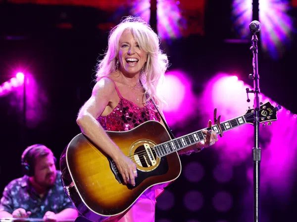Female country singer in pink dress playing acoustic guitar on stage with vibrant purple lighting and smiling joyfully.