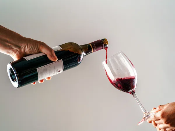 Red wine being poured from a bottle into a glass held by two hands against a plain background.