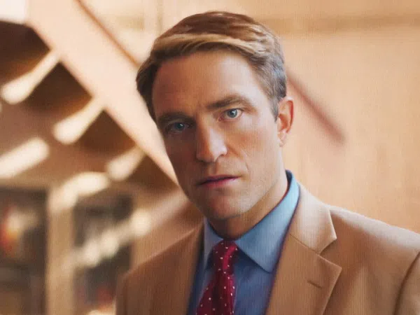 Man in a tan suit and red polka dot tie staring directly at the camera with a blurred warm background.