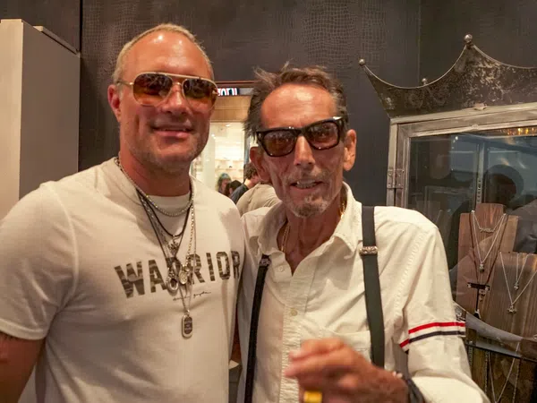 Two men wearing sunglasses and necklaces posing indoors at a jewelry store with display cases behind them