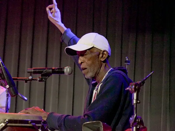 Elderly male musician playing congas and singing into a microphone with one hand raised on stage.