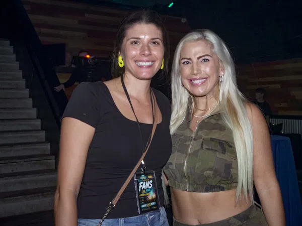 Two smiling women posing indoors, one in black top and the other in camouflage crop top with long blonde hair.