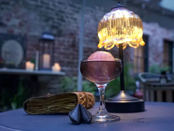 Elegant cocktail glass with a pink spherical garnish on a table beside a decorative lamp and fortune cookies.