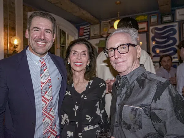 Three adults smiling together indoors, dressed in semi-formal attire at a social gathering with framed art on walls.