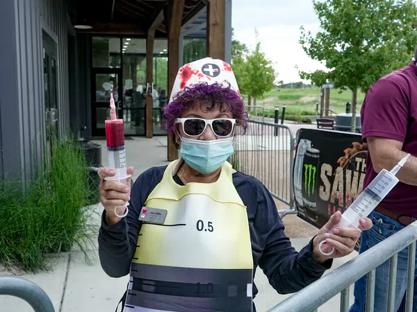 Person in nurse costume wearing sunglasses and mask holding two large syringes outside a building.