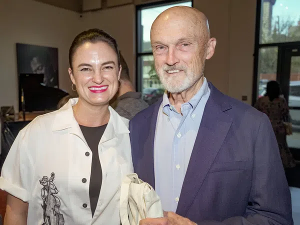 Smiling woman and elderly man standing indoors at a social event with large windows in the background.