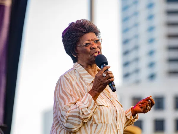 Elderly woman in striped blouse speaking into microphone at outdoor event with city building background