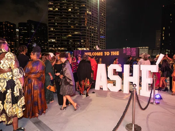 People dressed in elegant attire gather at a nighttime outdoor event with large illuminated ASHE letters and city buildings in the background