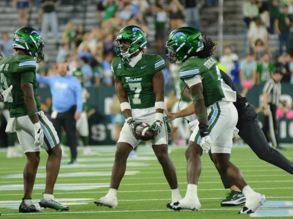 Tulane football players in green uniforms huddle on field during game with crowd in background