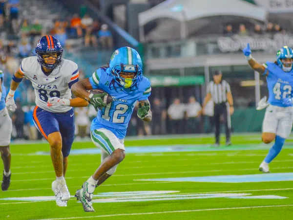 Football player in blue uniform running with ball as opponent in white uniform chases him on the field.