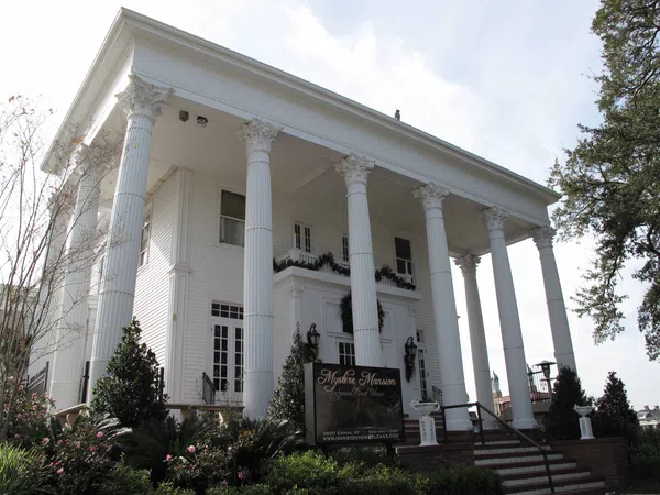 White Greek Revival mansion with tall Corinthian columns and manicured garden in front under a clear sky.