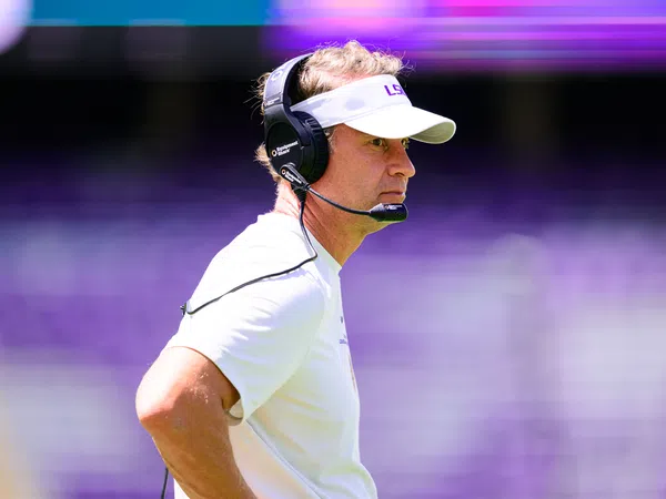 Football coach wearing LSU visor and headset focused during a game with blurred purple stadium background