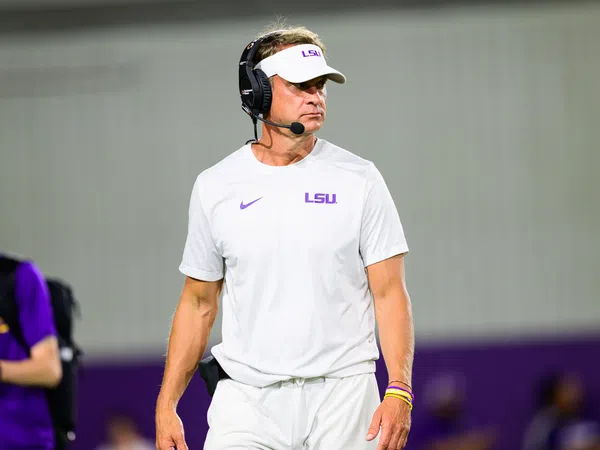 LSU football coach in white LSU gear and visor wears a headset on the sidelines during a game.