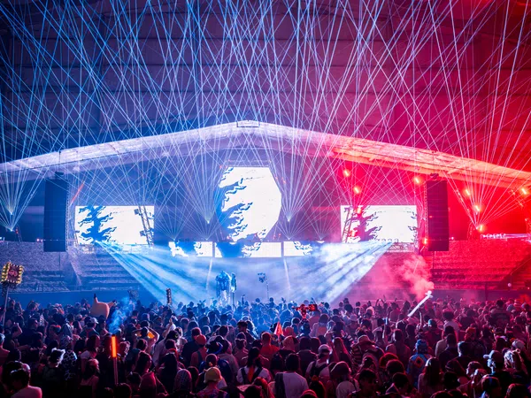 Crowd enjoying a nighttime concert with vibrant laser lights and colorful stage effects under a large canopy.