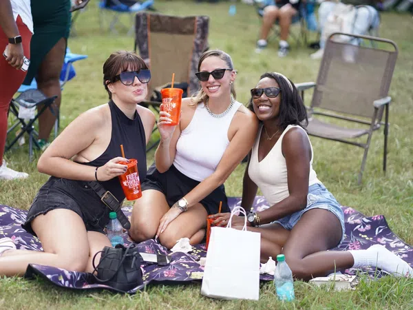 Three women wearing sunglasses enjoying drinks while sitting on a blanket at an outdoor event on grass.