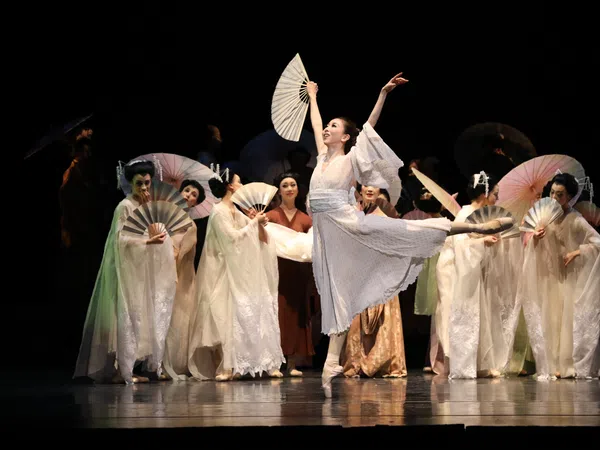 Ballet dancer in white costume performs with fans on stage with other dancers in traditional attire.