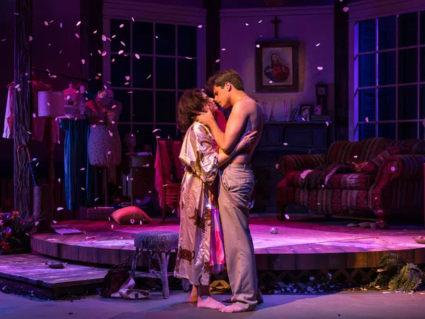 Couple embraces on stage in intimate theater scene with warm lighting and scattered petals.