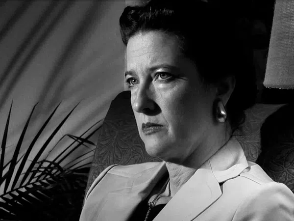 Black and white portrait of a serious woman sitting in a chair with shadows and a lamp in the background