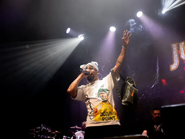 Hip hop artist performing on stage under bright spotlights with microphone and raised arm during concert.