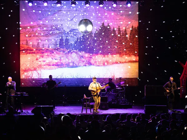 Band performing on stage with snow effects and forest landscape backdrop under disco ball lighting.