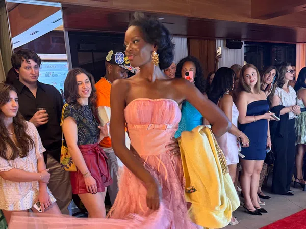 Model in pink gown holding yellow jacket walking past crowd of spectators at indoor event with wood paneling.