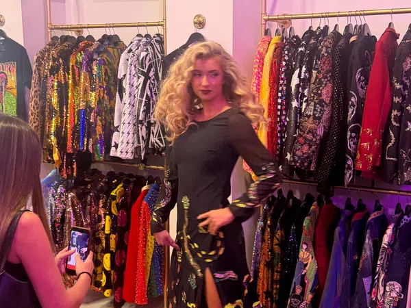 Woman in black dress posing in a colorful boutique with sequined and patterned jackets on racks