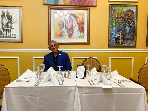 Man in blue suit sitting at white tablecloth-covered table set with wine glasses and silverware, framed art on yellow wall.